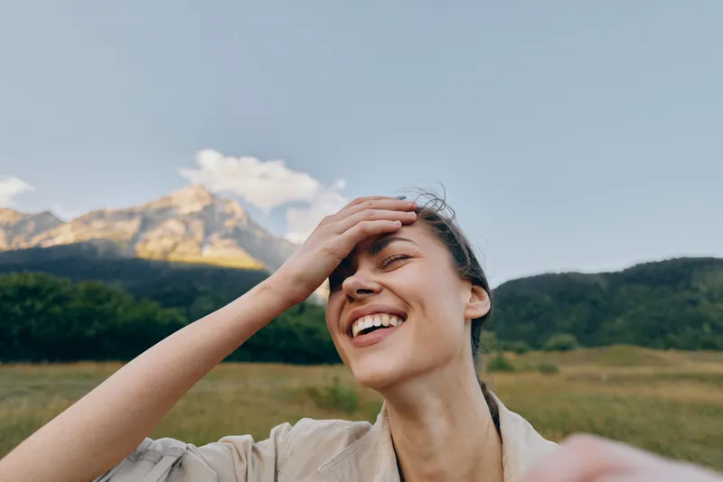 Woman enjoying outdoor lifestyle, smiling and carefree, standing in a sunlit field with distant mountains, bright sky, capturing a joyful nature moment in open countryside