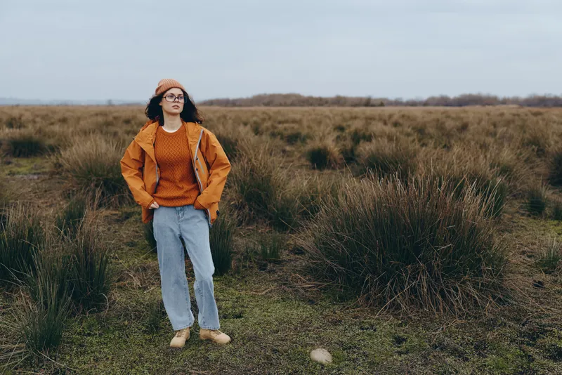 Woman outdoors walking through a grassy field, casual lifestyle, wearing hat and sweater, jeans and boots, relaxed smile, soft daylight, natural grassland