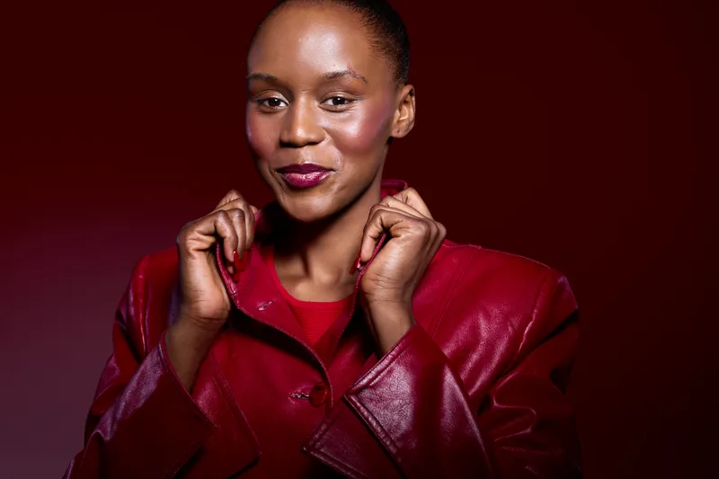 Confident dark skinned African woman wearing a red leather jacket smiles softly against a dark red background. Concept of fashion, style, beauty, and elegance with natural makeup.