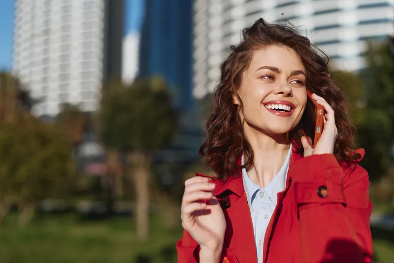 Cheerful woman outdoors, smiling while talking on the phone, wearing a red jacket, sunlit city background, relaxed mood, natural colors, candid moment