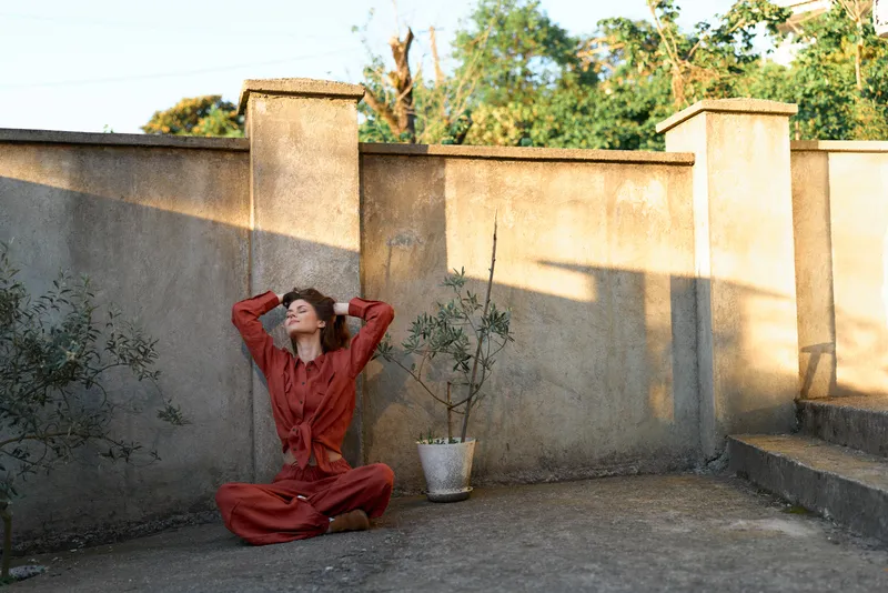 Woman wearing red dress sits on concrete ground in an outdoor courtyard, leaning back with hands behind head, serene lifestyle moment captured against a warm wall