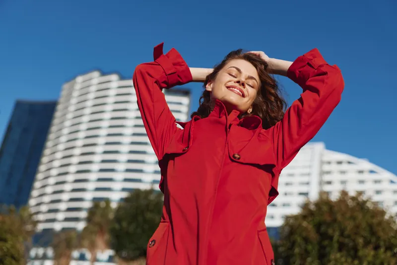 A happy woman in a red coat enjoys sunlight outdoors with eyes closed and hands behind head. Modern white building and blue sky are visible in the background.