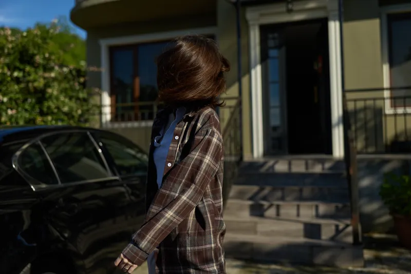 Woman lifestyle scene: a woman walks outside a modern house in plaid coat, casual outfit, sunny day, relaxed mood, candid everyday moment