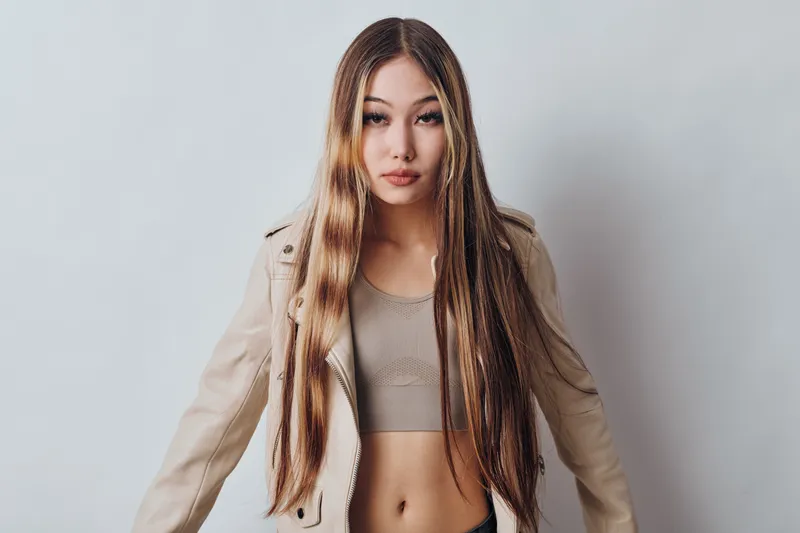 Young woman with long straight hair wearing beige cropped top and light jacket standing against plain white background looking at camera with neutral expression.