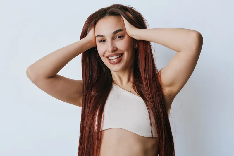 Young woman with long red hair smiling and posing with hands on head against light background. Casual style, happy expression, natural makeup and relaxed posture in studio.