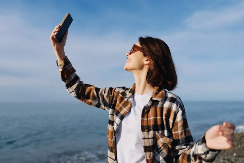 Woman taking a selfie by the sea in a plaid shirt, outdoors, casual style, holding a phone toward the horizon, sunny day, relaxed mood and travel vibe.