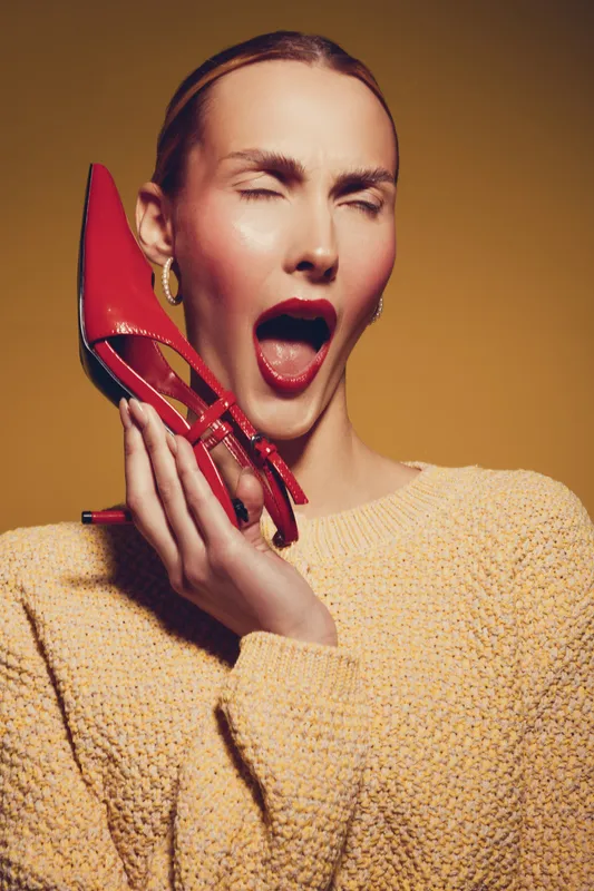 Young woman in yellow sweater holds red high heel shoe near face, eyes closed and mouth open. Studio portrait with warm background. Concept for fashion, style, and emotions.