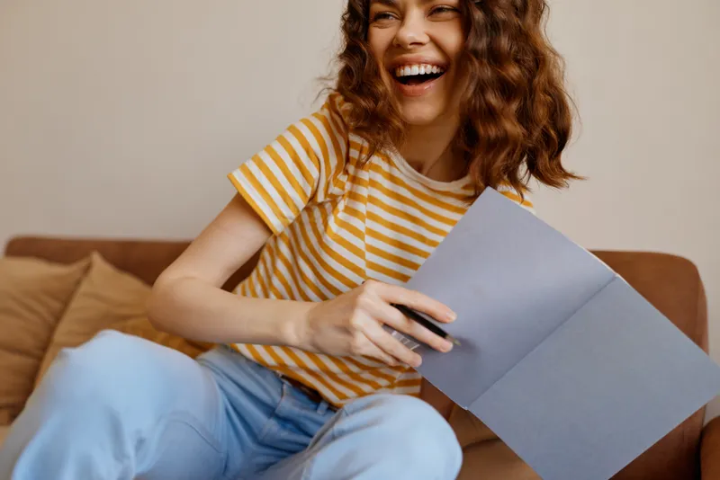 Woman enjoying a casual home moment, smiling while holding a card and envelope on a sofa, striped yellow shirt, relaxed lifestyle scene, cozy interior.