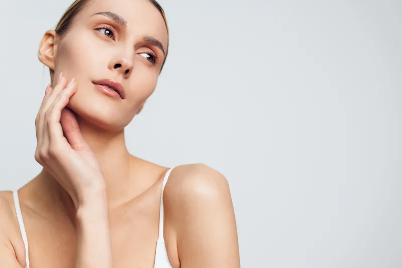A young woman with clear skin gently touches her face while looking away thoughtfully against a plain light background. Concept for skincare, beauty, and natural health.