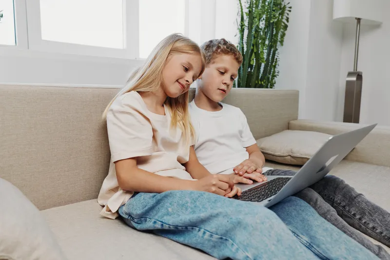 Two children sitting on a sofa using a laptop together in a bright living room. Casual clothing and relaxed atmosphere emphasize family and technology interaction.