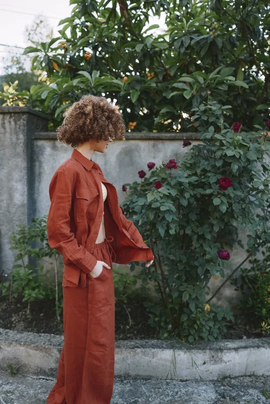 Young woman with curly brown hair walks beside a rose garden in a rust orange jacket and matching pants, radiating casual confidence, with soft sunlight and a relaxed mood