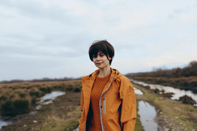 Woman in orange jacket on outdoor path in countryside, nature portrait, walking stance, looking at camera, casual lifestyle outdoors, cloudy sky, calm spring atmosphere