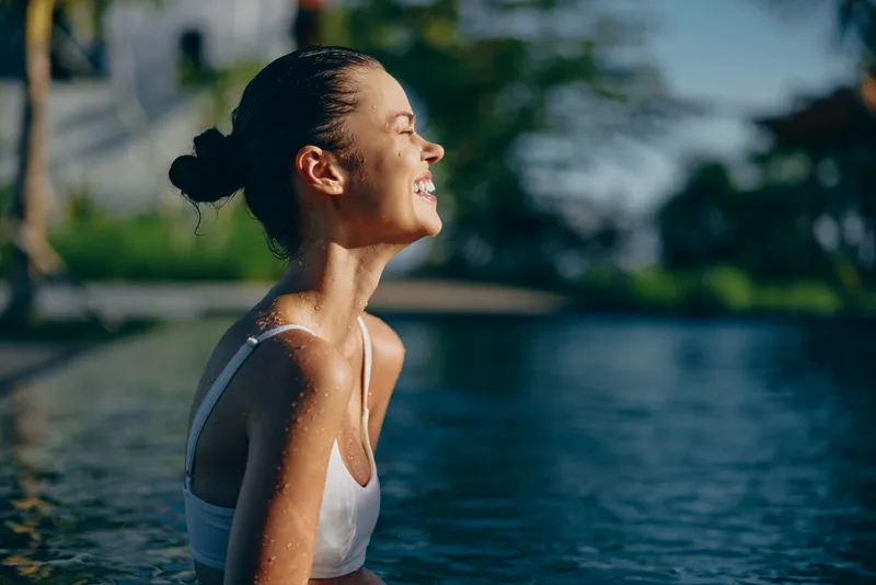 Young woman in white swimsuit enjoying sunlight while sitting in outdoor pool. Relaxed lifestyle concept with natural background and warm summer atmosphere.