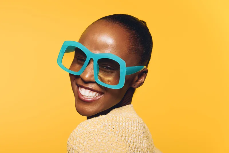 Happy dark skinned African woman smiles wearing large blue sunglasses and beige sweater, posing on yellow background. Concept of joy, fashion, summer style, and confident beauty portrait.