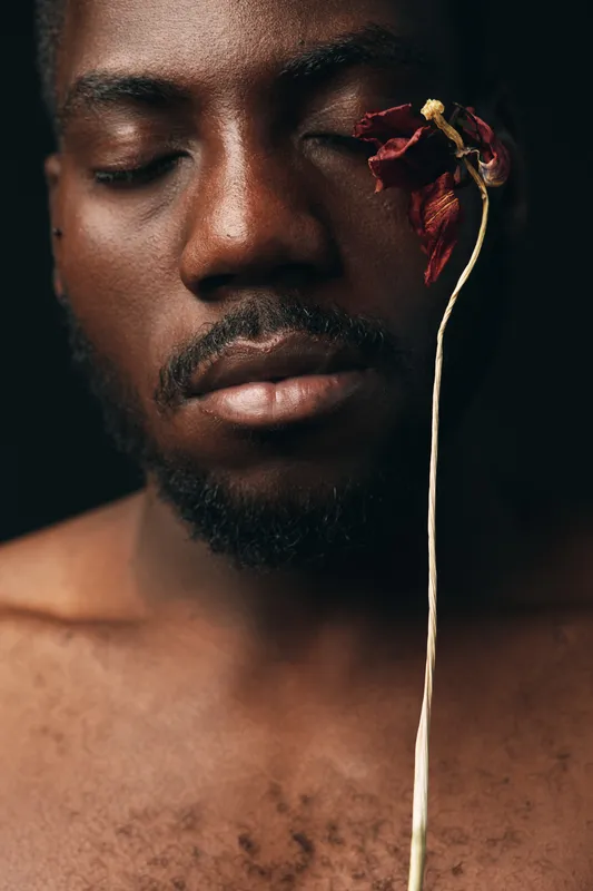African man with closed eyes holding dried flower near face in creative conceptual visual style. Portrait of dark skin male with beard and artistic expression on black background.