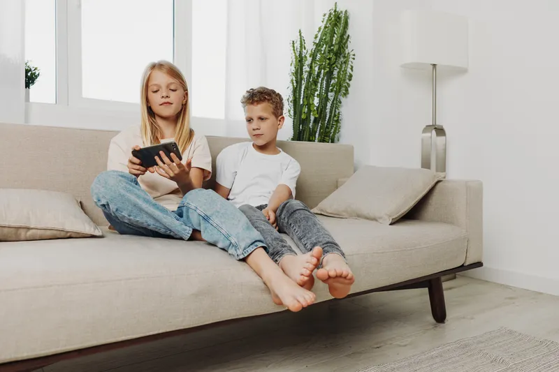 Two children sitting barefoot on sofa indoors, girl holding tablet, boy watching screen attentively in bright modern living room.