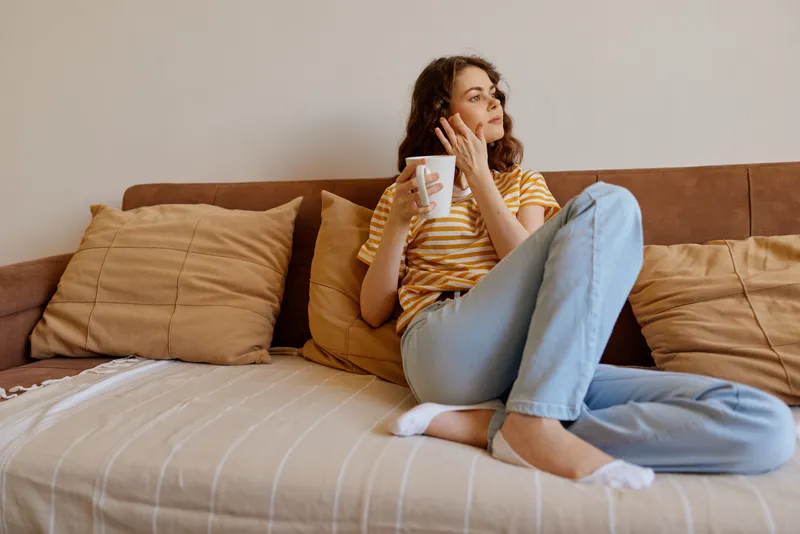 Cheerful woman at home, relaxing on a sofa, striped shirt, jeans, holding a cup, natural light, casual lifestyle, cozy living room, candid moment, everyday comfort