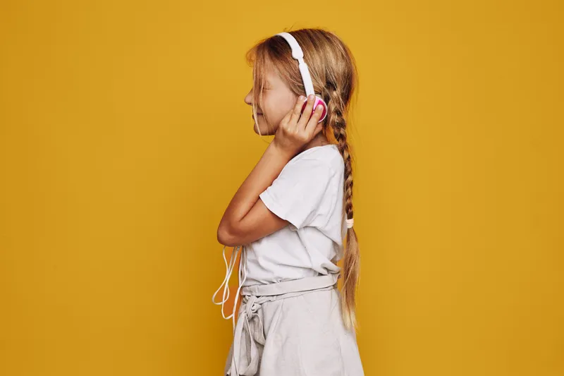 Young girl with braided hair listens to music with white headphones. She wears a white t-shirt and light pants against a solid yellow background. Side view portrait.