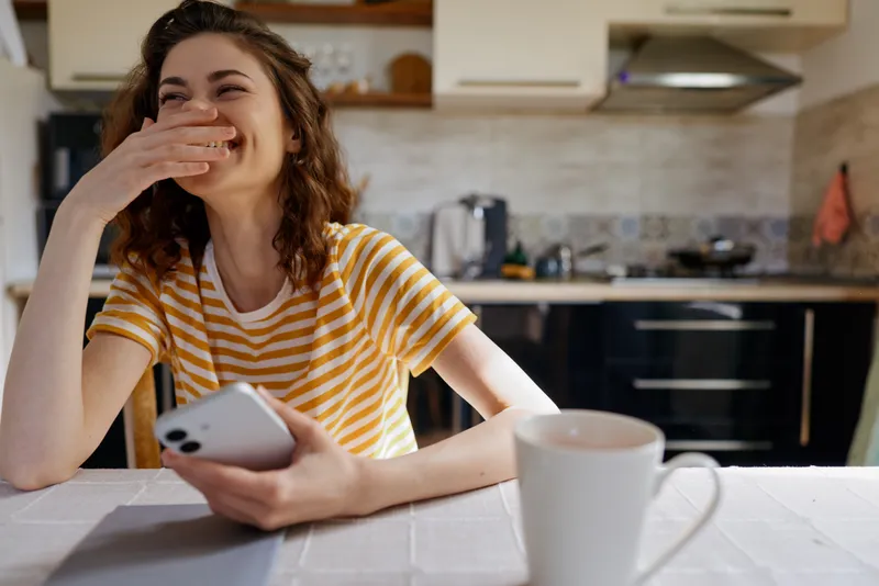 Young woman in striped tshirt sits at a bright kitchen table, laughing with her hand over her mouth while scrolling on her phone, capturing a candid home moment.