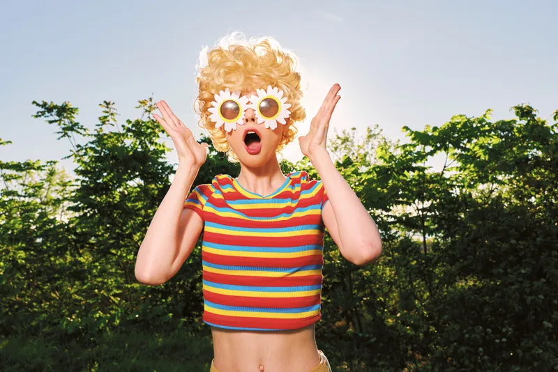 Cheerful woman outdoors, wearing a striped crop top and playful flower sunglasses, surprised expression, sunlit lawn, retro film color, vibrant greens, candid lifestyle moment