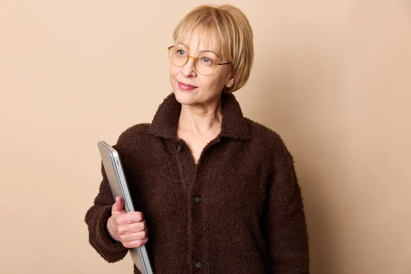 Elderly woman with glasses smiling in studio portrait holding tablet, looking at screen, using digital technology for learning and online communication on neutral beige background