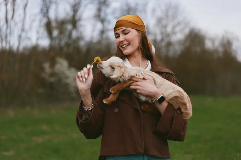 A joyful woman outdoors shares a gentle moment with a young goat, smiling as she offers a dandelion, creating a warm, natural lifestyle scene of affection between human and animal.