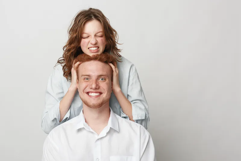 Playful young woman with curly hair squeezes head of smiling redhead man in white shirt. Studio portrait on light background. Concept of friendship, fun, humor, and casual interaction between friends.