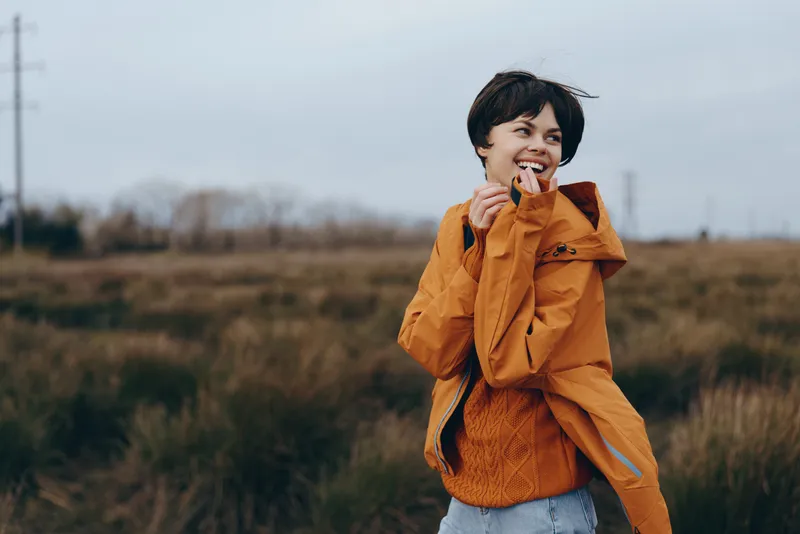 Woman lifestyle, outdoors, smiling, orange, jacket, field, casual, spring breeze, woman in rural meadow, hand near lips, warm jacket over muted grass, soft overcast light, natural colors, relaxed