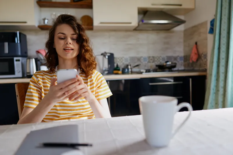 Young woman in a striped yellow tee sits at a bright kitchen table, smiling as she looks at her smartphone, with warm natural light and a casual, relaxed mood.