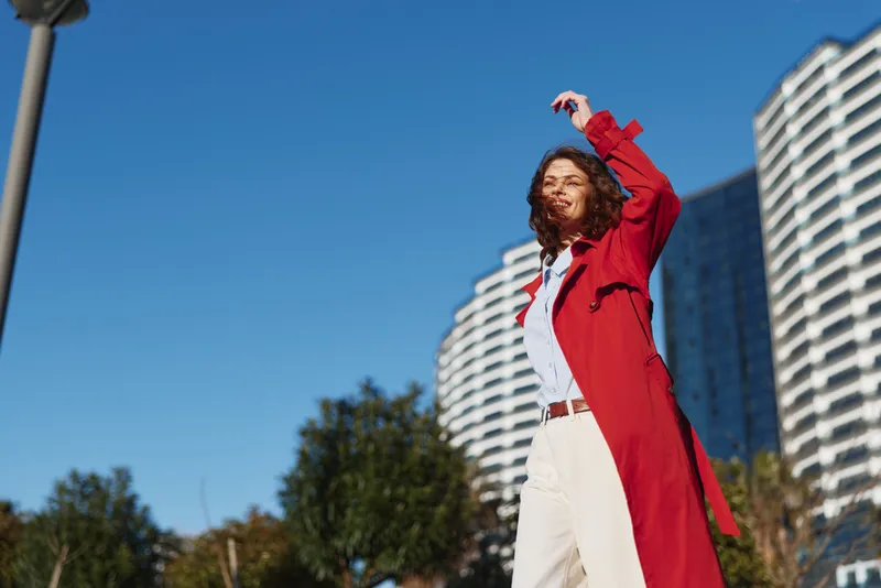 A young man with curly hair in a red coat and white pants stands outdoors in an urban park, stretching his arms toward a clear blue sky, embodying bold confidence and freedom.