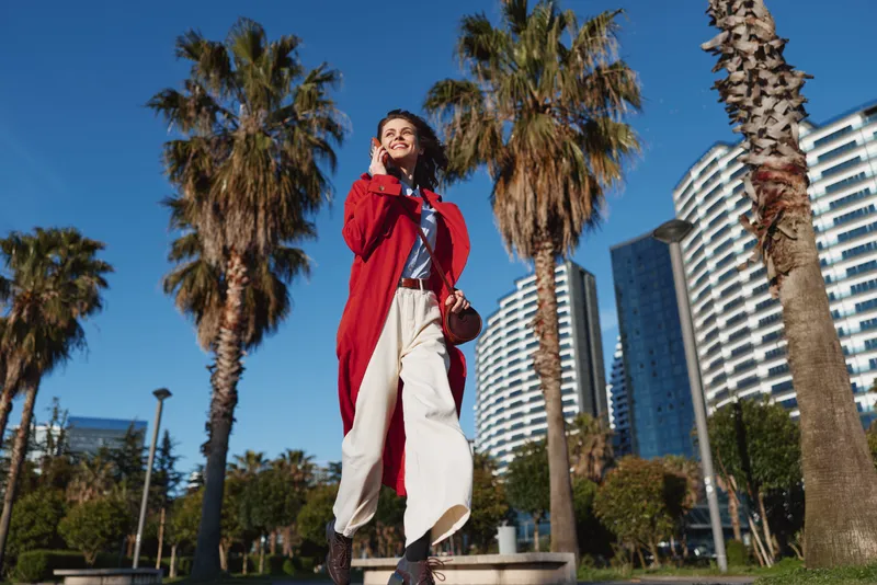 Young woman in a red coat stands under palm trees in a sunny city, talking on her phone with a confident smile, breeze in her hair.