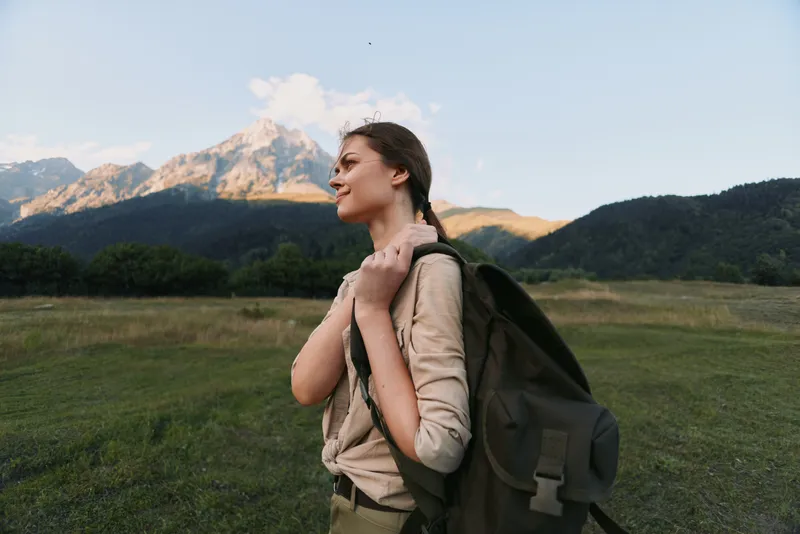 Young woman with a backpack stands in a sunlit meadow, gazing toward distant mountains, enjoying a peaceful outdoor moment in a light breeze for fresh air and freedom