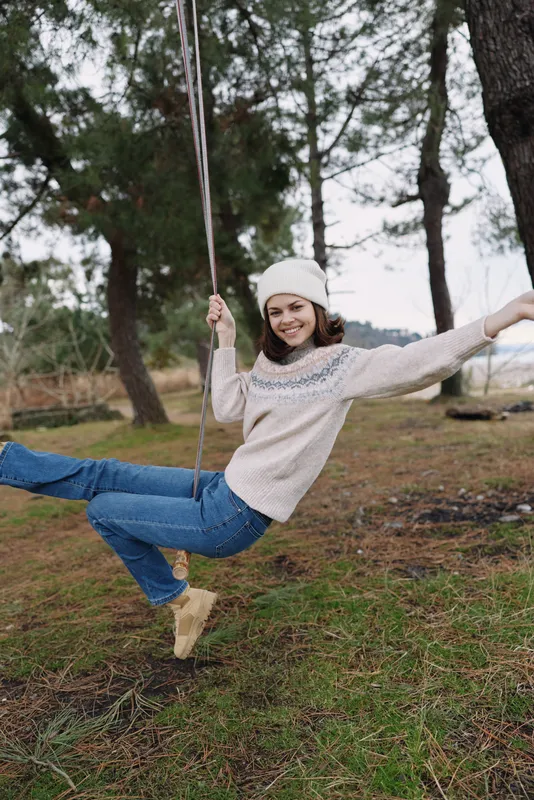 Girl on a swing in an outdoor park, winter scene, wearing a sweater and beanie, smiling and enjoying movement, casual playful moment in nature.