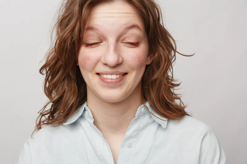 Young woman with curly hair smiles with closed eyes wearing light shirt on white background. Concept of natural beauty, happiness, and relaxed positive mood in casual portrait.