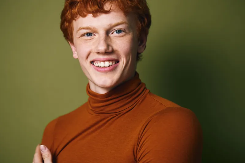 Young man with red hair smiling wearing a brown turtleneck sweater against green background in portrait photography studio.