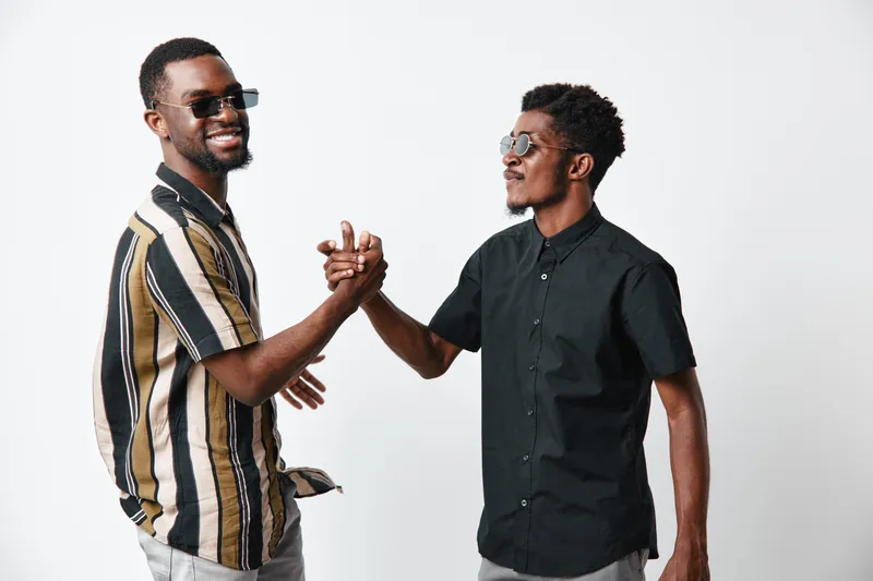 Two African American men with dark skin shaking hands and smiling in casual clothes and sunglasses on white background for friendship and connection concept.