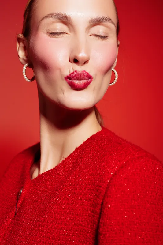 Close-up of a woman with red lipstick and earrings making a kiss face. She wears a red textured top against a red background. Concept of beauty, makeup, and fashion.