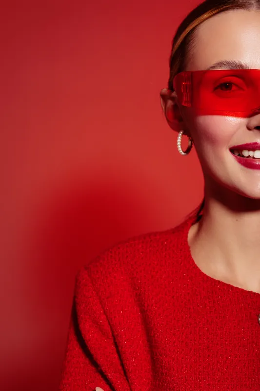 Smiling woman in red sweater and red sunglasses poses against red background. Close-up of happy female model with hoop earrings and bright makeup. Concept of fashion and style.