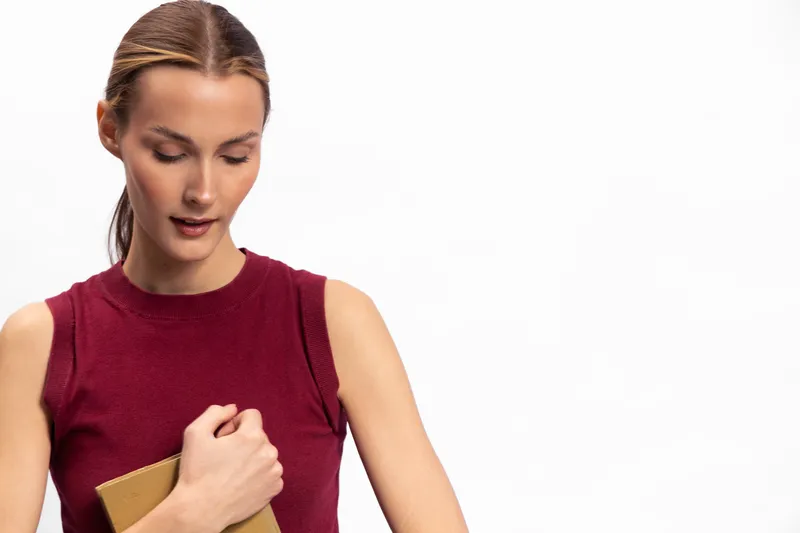 Young woman in sleeveless red top holds closed book close to chest, looking down thoughtfully. Minimalist white background highlights calm and introspective mood of the portrait.