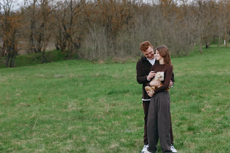 Couple outdoors in a grassy park cradle a small goat kid, smiling and enjoying a gentle moment. Warm natural light emphasizes tenderness, care, and family life in a simple rustic setting.