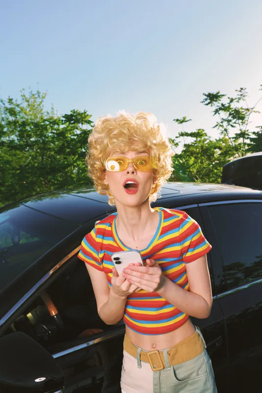 A surprised woman with curly blonde hair in a striped rainbow top stands beside a car, phone in hand, yellow sunglasses, bright retro hues, outdoor sunlight and casual lifestyle.