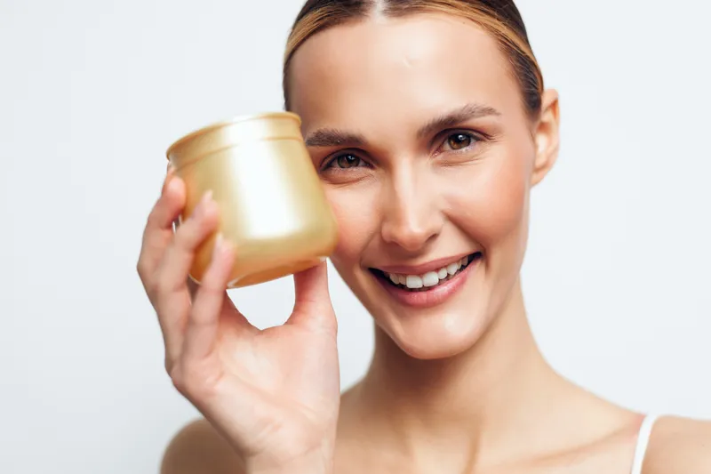 Smiling woman holds golden cosmetic jar close to face shows skincare product with clean fresh skin and natural makeup isolated on white background beauty and wellness concept.