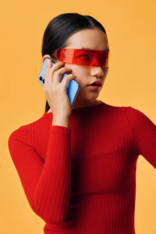 Woman in red sweater and futuristic red visor talking on smartphone against yellow background. Modern style, technology use, communication and fashion portrait.