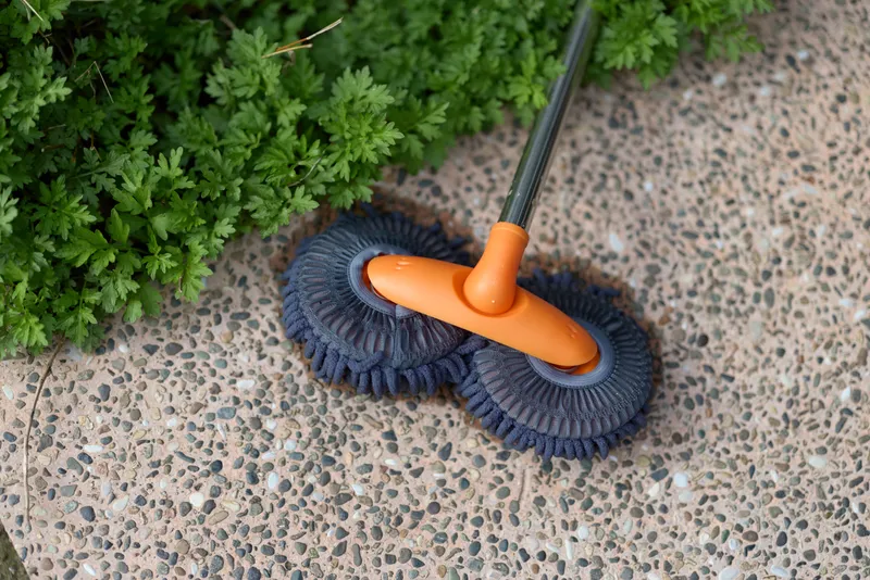 Close-up of a two-tone cleaning brush with an orange handle and blue bristles on a speckled terrazzo floor beside a green plant, ready for a quick indoor cleaning.