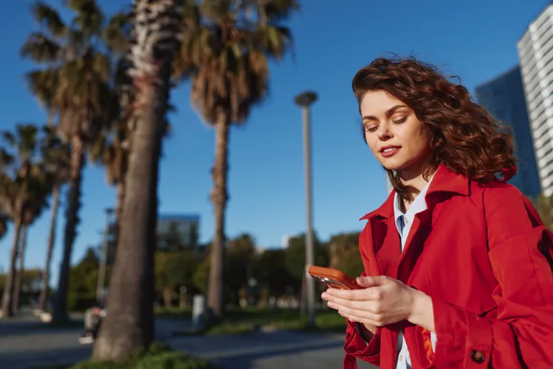 Young woman in a redcoat checks her smartphone outdoors with palm trees and a cityscape behind, a bright windy moment conveying focus and urban energy.