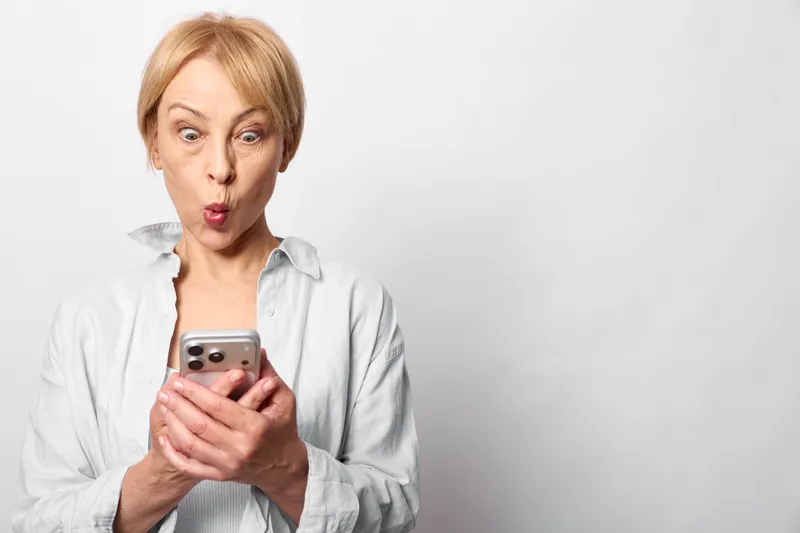 Woman smartphone surprised shocked communication technology portrait indoors white background, female blond short hair holding mobile phone, open mouth reaction, copy space