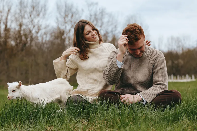 Young couple in cozy sweaters relaxes on grass near a white baby goat in a natural outdoor setting during a cool autumn day.