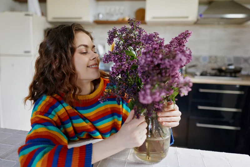 Cheerful woman in a colorful striped sweater admires a bouquet of lilacs in a cozy kitchen, capturing a warm everyday home moment with natural light and a soft smile.