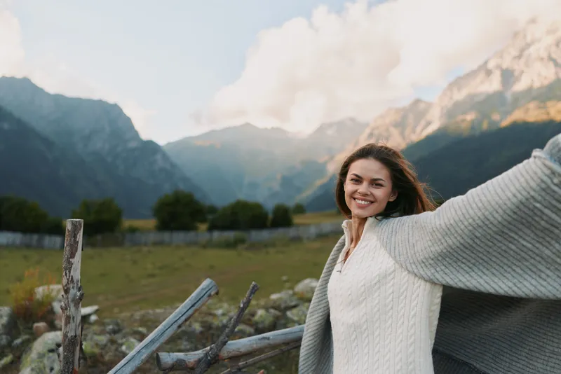 A smiling woman stands outdoors in a mountain landscape, wearing a light sweater and a flowing cape, arms open wide. Concept for freedom, wellbeing, and outdoor lifestyle.