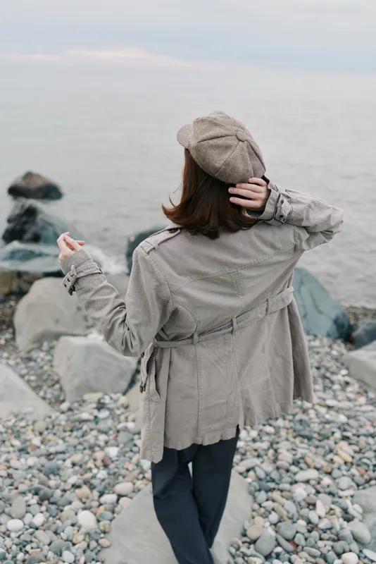Woman wearing beige coat and matching beret stands on rocky beach looking at calm sea. Concept of autumn fashion, solitude, and peaceful coastal environment.